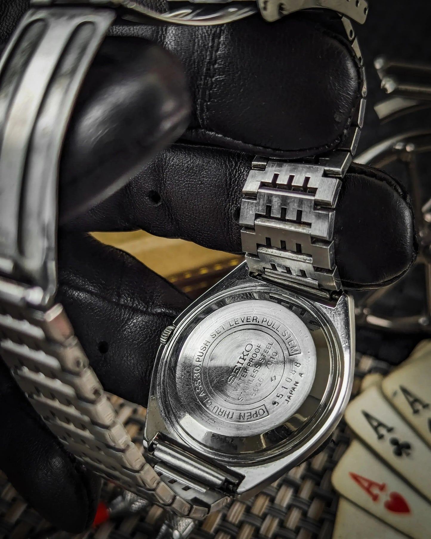 Close-up of Seiko stainless steel watch case back held with black gloves, playing cards in background