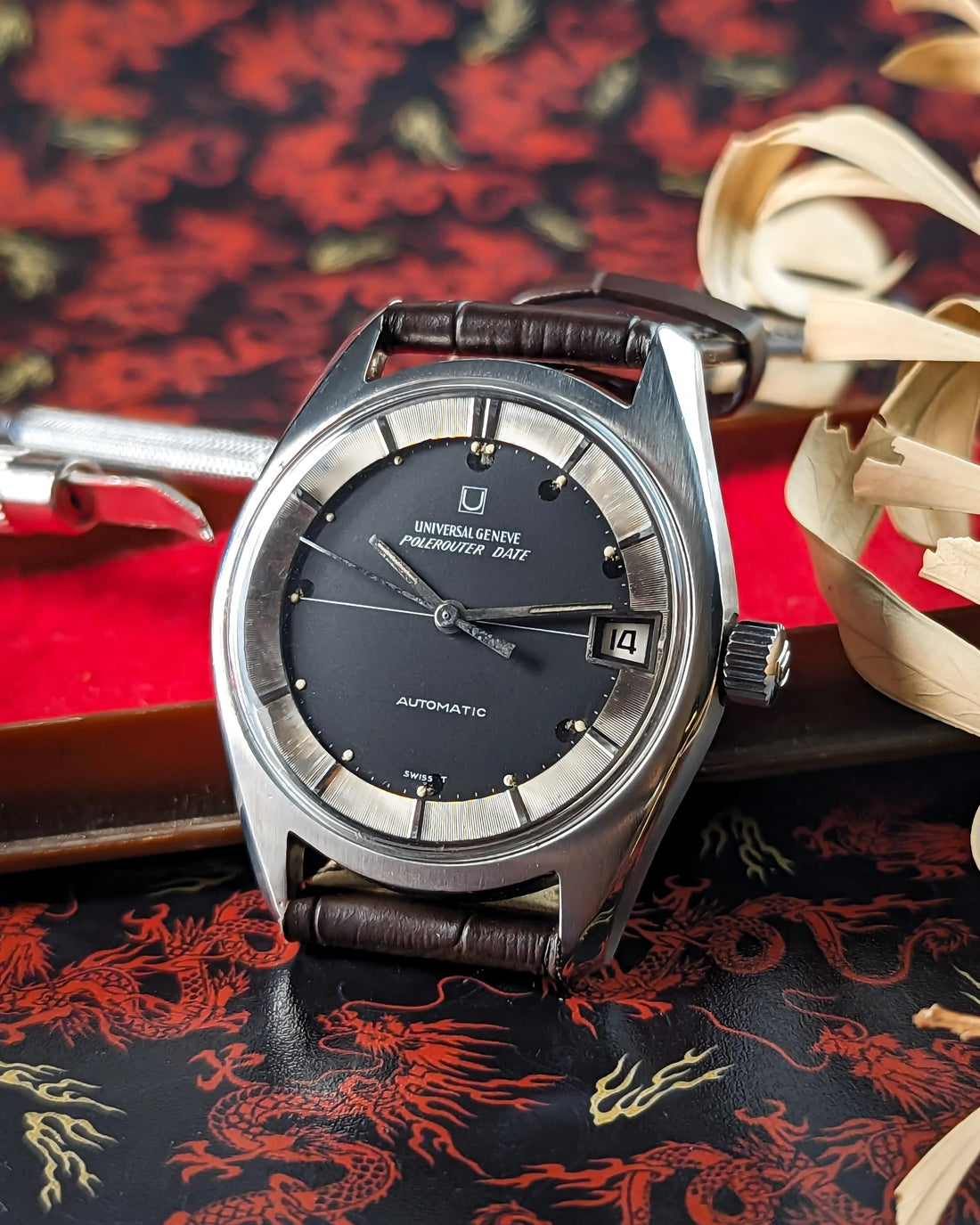 Classic Universal Geneve Polerouter watch with black dial, silver markers, brown leather strap on ornate dragon-patterned background.
